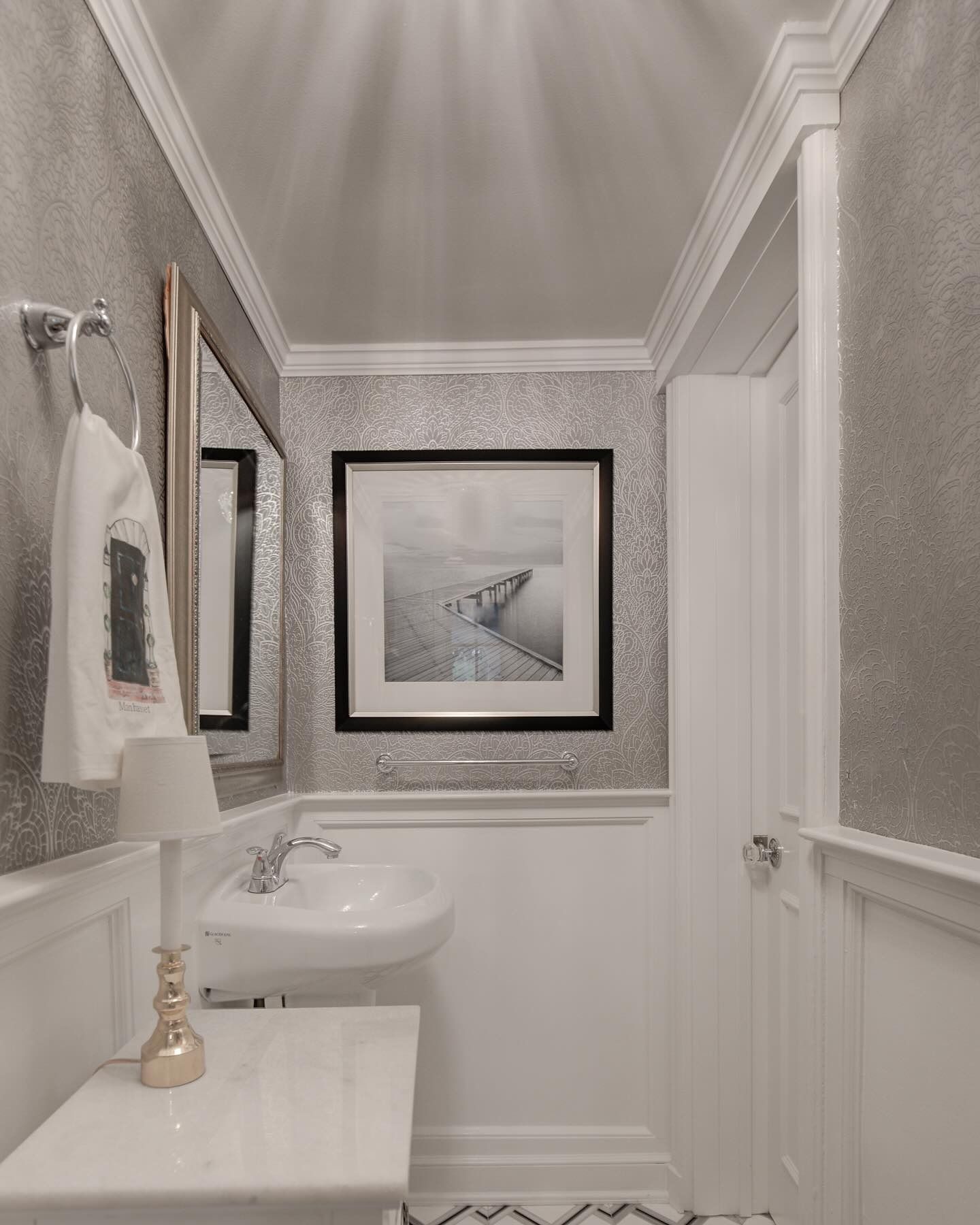 Small powder room with silver wallpaper, white wainscoting, a sink, and a framed grayscale photo.