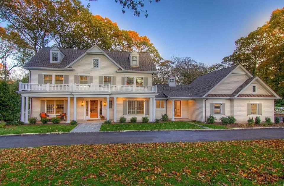 Two-story white house with light gray shutters, porch, and a driveway lined with fallen leaves.