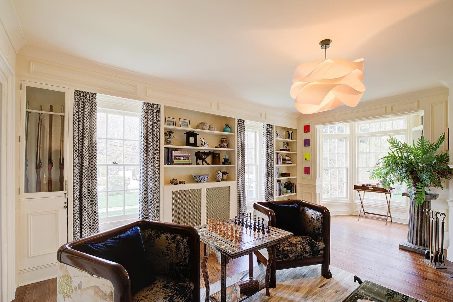 Living room with built-in bookshelves, two armchairs facing a chess board, and large windows with patterned curtains.