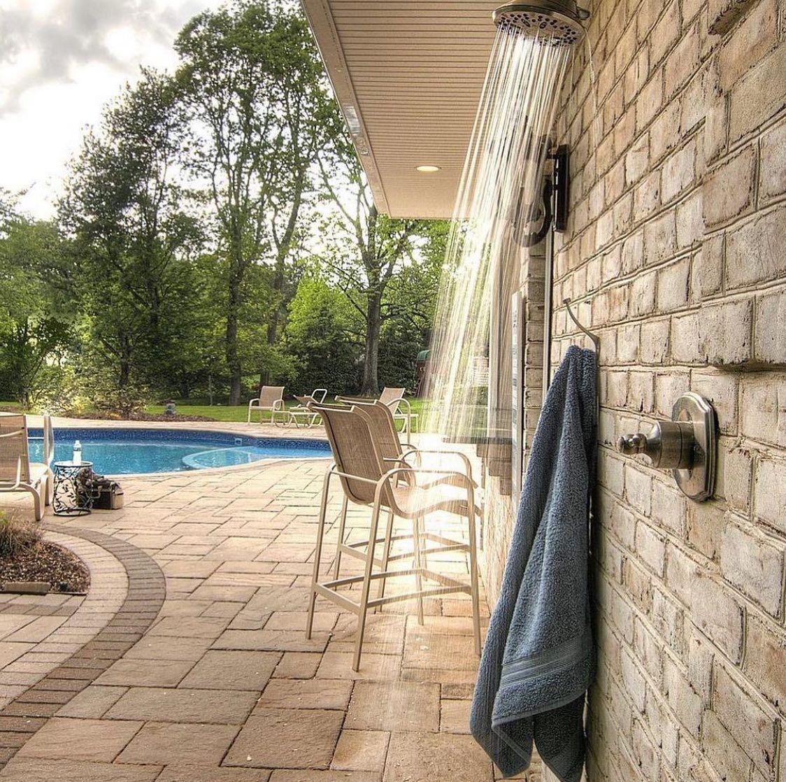 Outdoor shower next to brick wall. A blue towel hangs on hook. Pool and chairs in background.