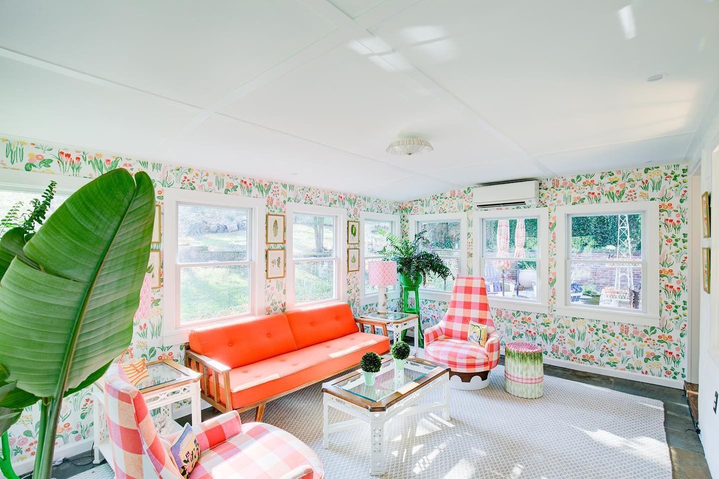 Sunroom with floral wallpaper, orange sofa, pink gingham chairs, and a white rug.