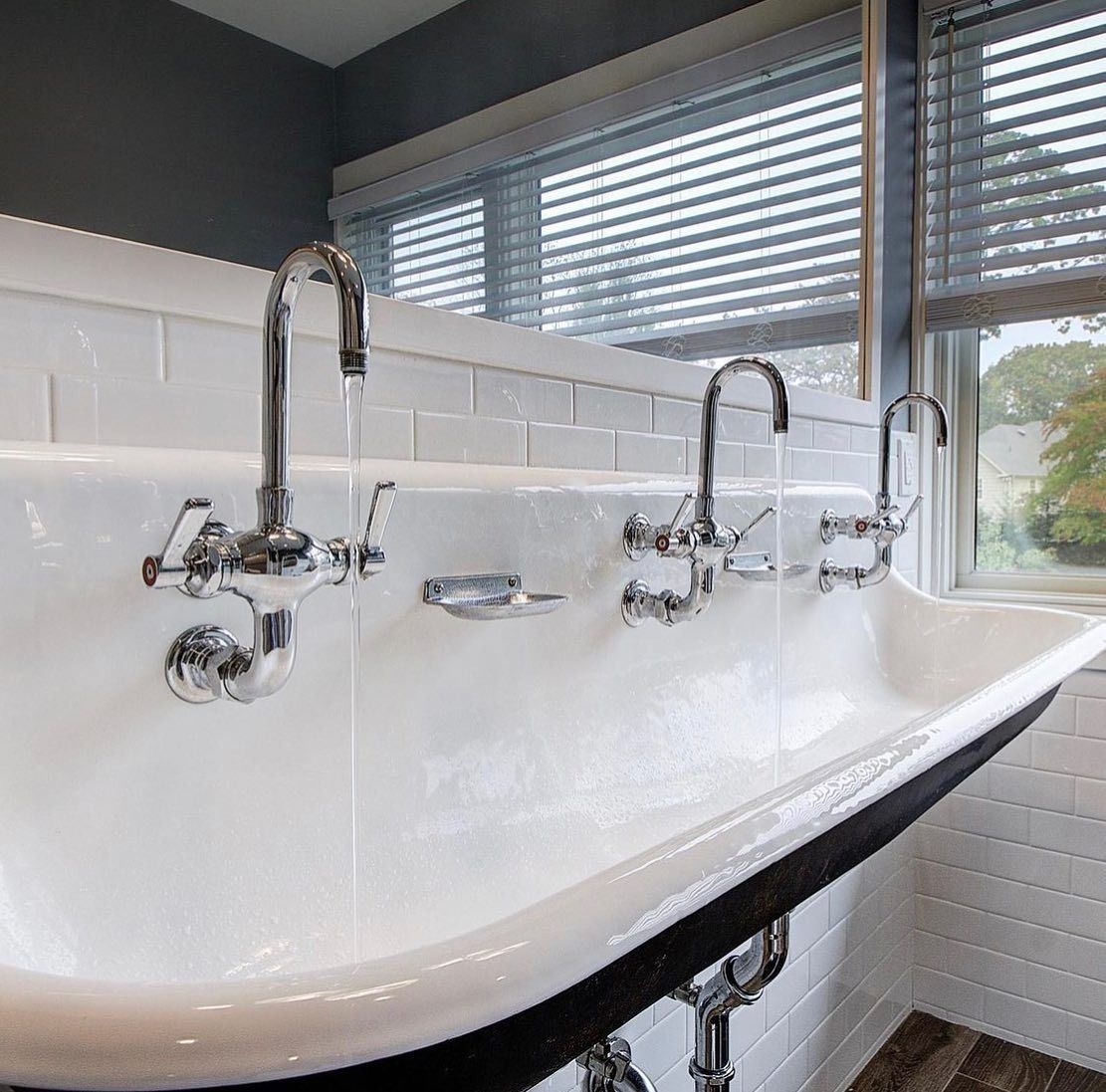 White, trough-style bathroom sink with three faucets; water running. Light coming through window.