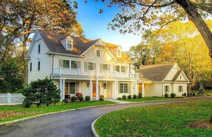 White colonial house with a long driveway and green lawn surrounded by trees.