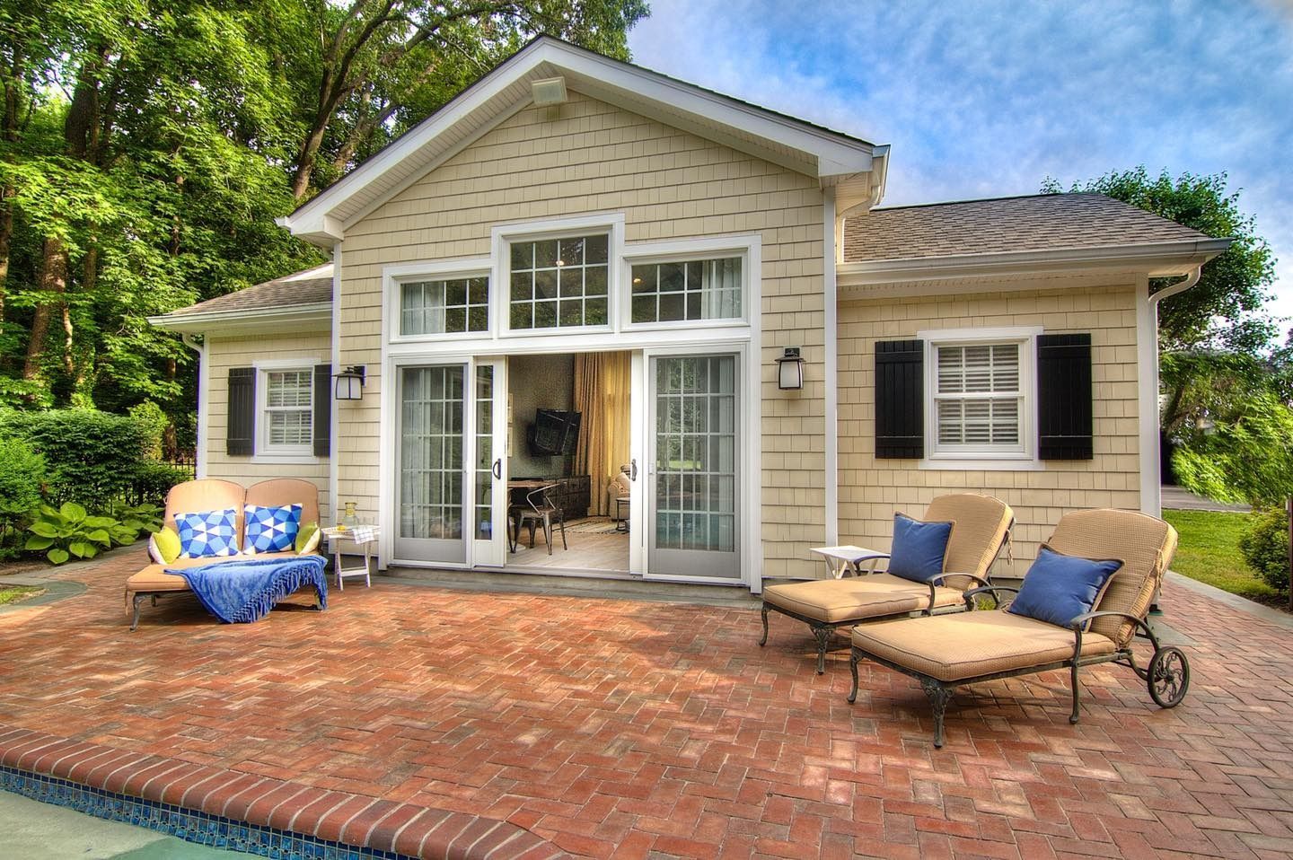 Beige cottage with brick patio, lounge chairs, and pool, surrounded by trees.