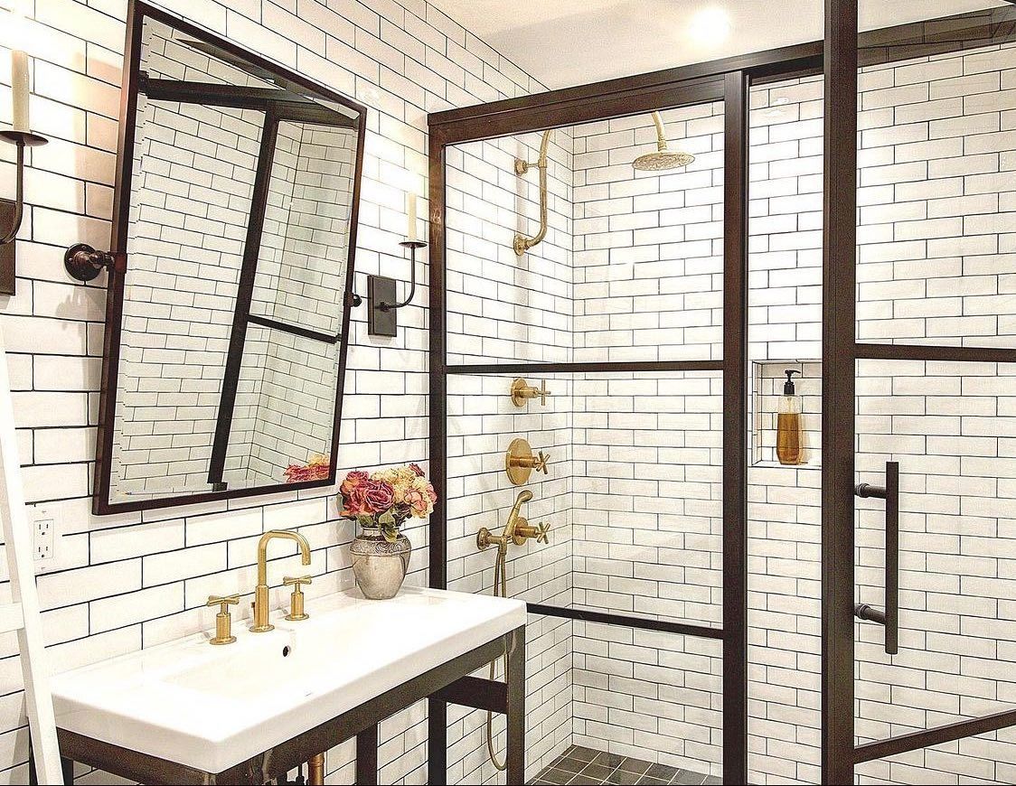 Bathroom with white brick tile, black-framed shower and mirror, gold fixtures, and a white sink.