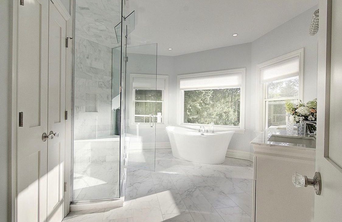 Bright white bathroom with marble floors, glass shower, and freestanding bathtub near windows.