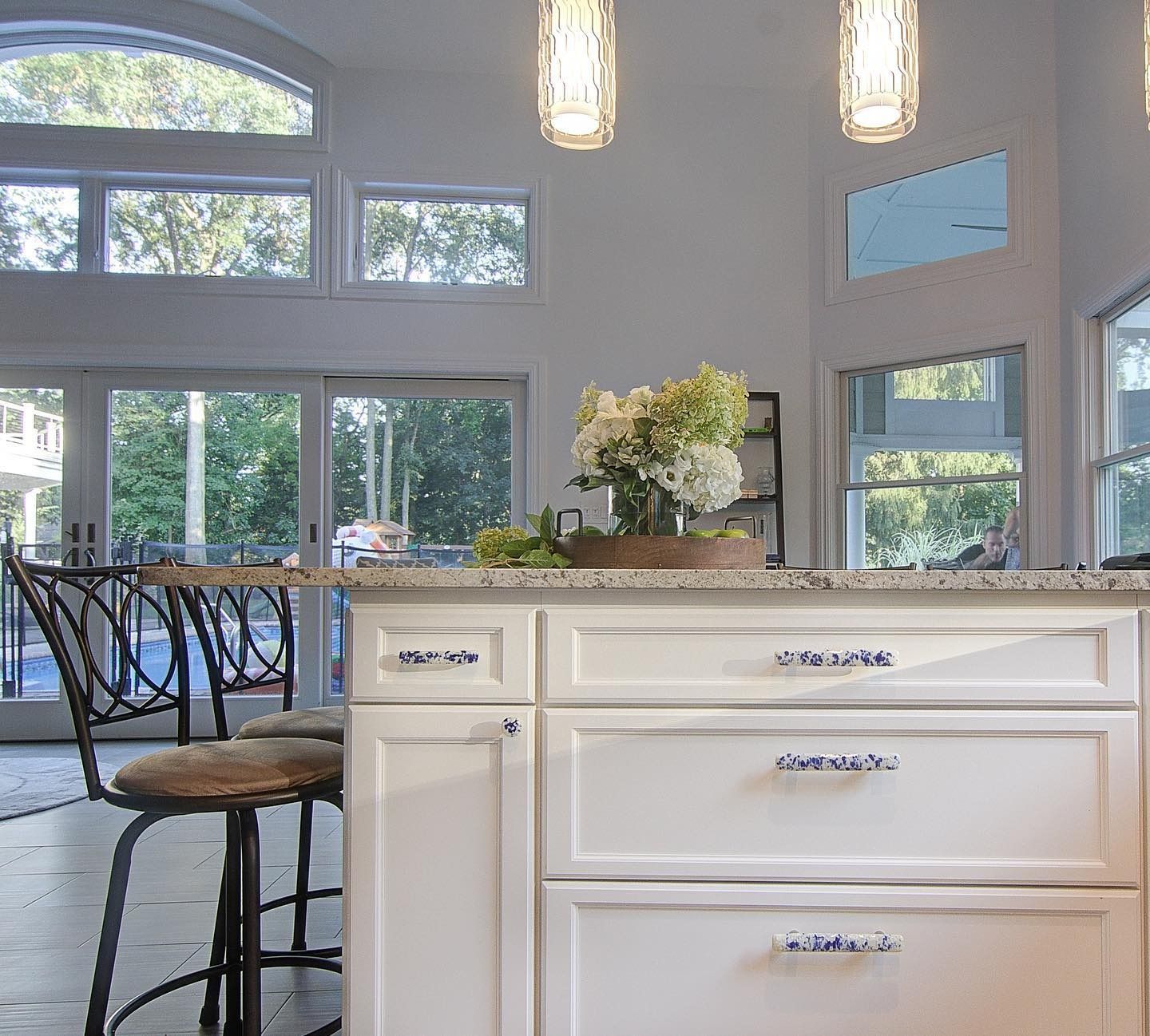 White kitchen island with drawers, flowers, and pendant lights; large windows in background.