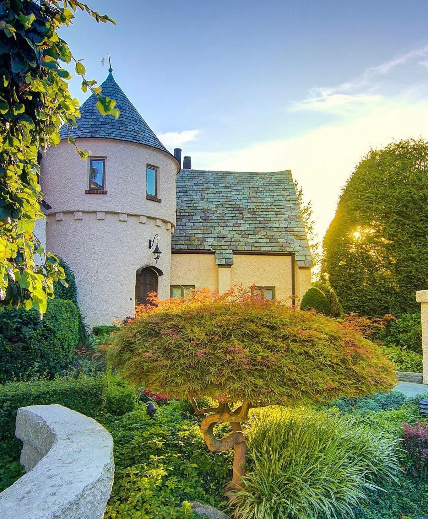 Stone castle with a turret and slate roof surrounded by lush green garden.