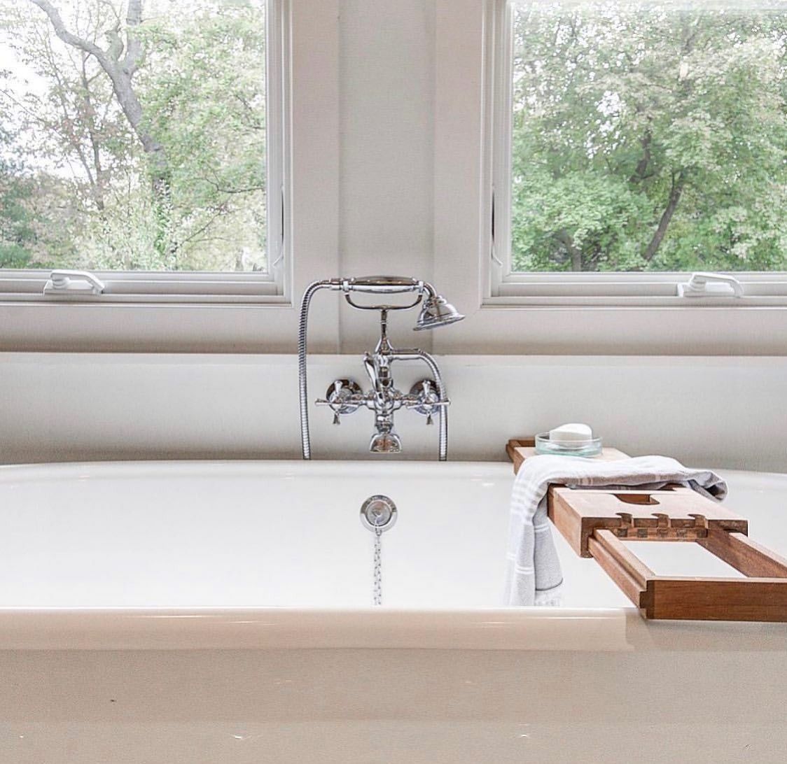 Bathtub near window, with bath caddy holding soap and towel. Chrome faucet and outdoor view visible.
