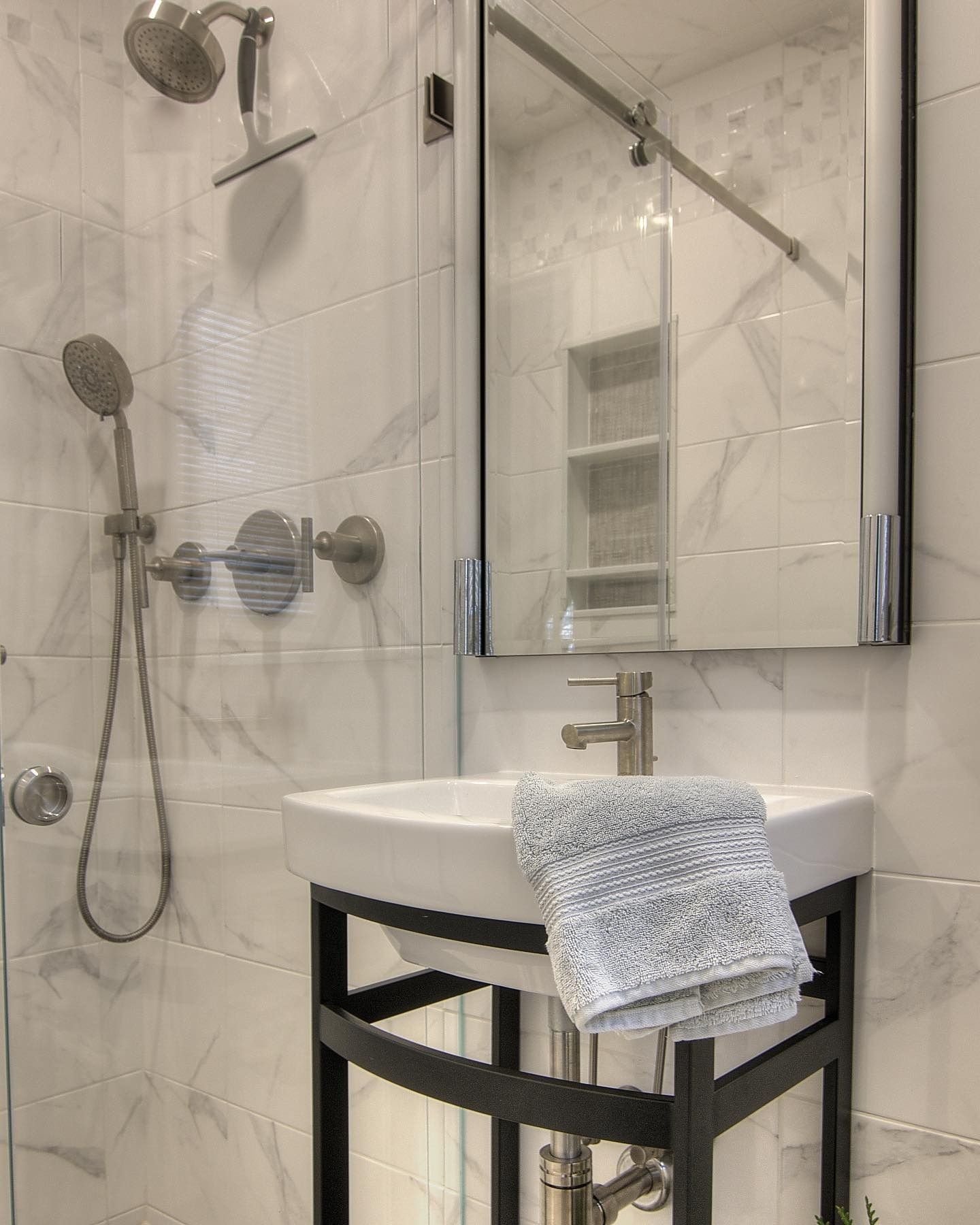 Small bathroom with white marble-look tiles, a square sink on a metal stand, and a framed mirror. A shower is to the left.