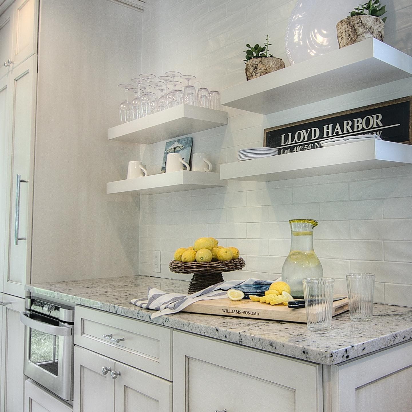 White kitchen with granite countertops, floating shelves, and a tray of lemons and glasses.