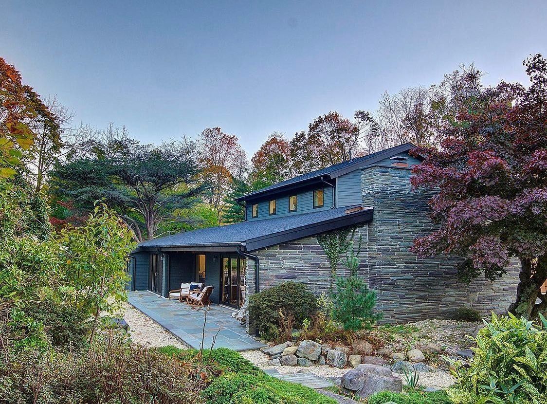 Stone house with a dark roof and a covered porch, surrounded by lush trees and greenery.