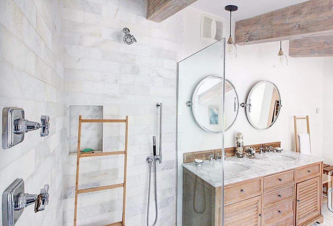 Bathroom with marble tile, wooden vanity, round mirrors, and glass shower door.