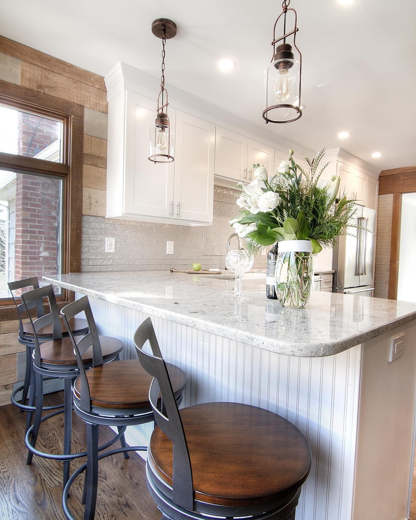 Kitchen with a white countertop island and white cabinets. Bar stools, pendant lights, and flowers are visible.