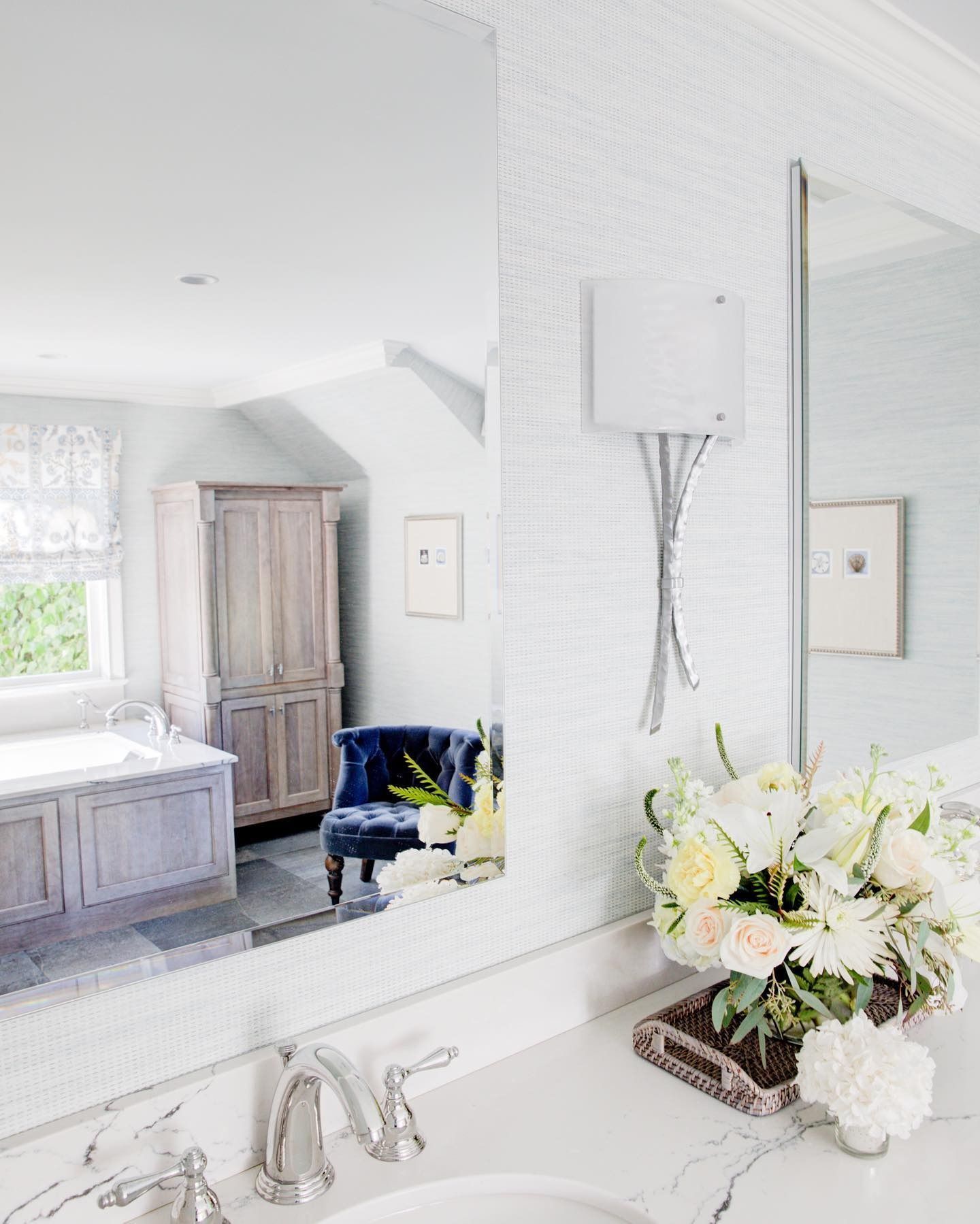 Elegant bathroom with large mirror, floral arrangement, blue accent chair, and light blue walls.
