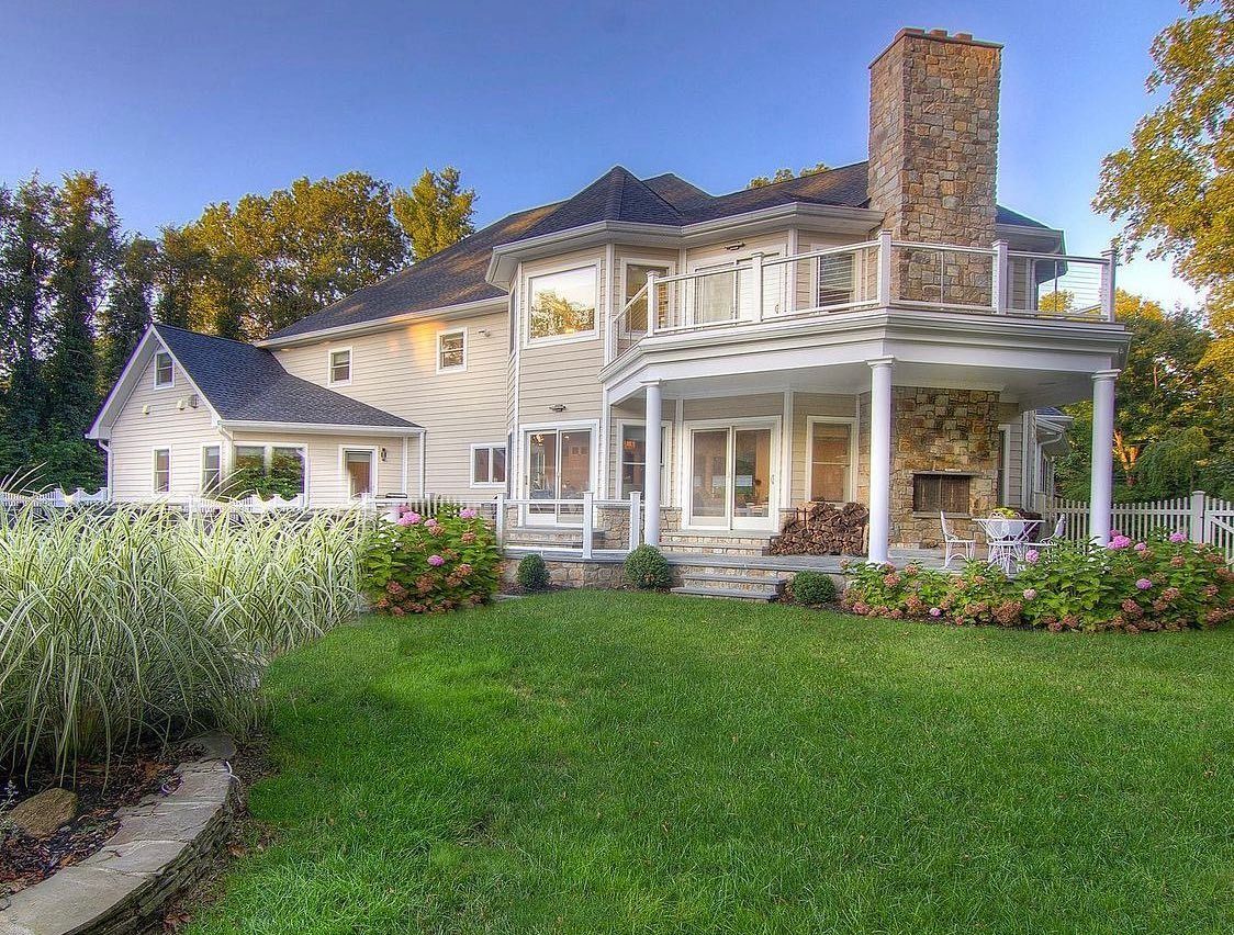 Two-story beige house with stone chimney and porch, surrounded by green lawn, bushes, and trees under a blue sky.