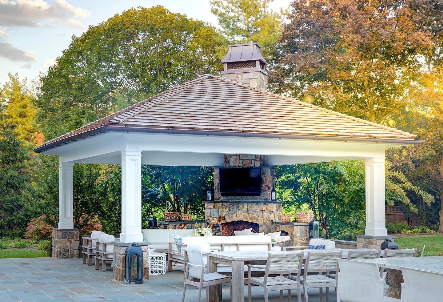 Outdoor gazebo with a fireplace, TV, seating, and a stone chimney.