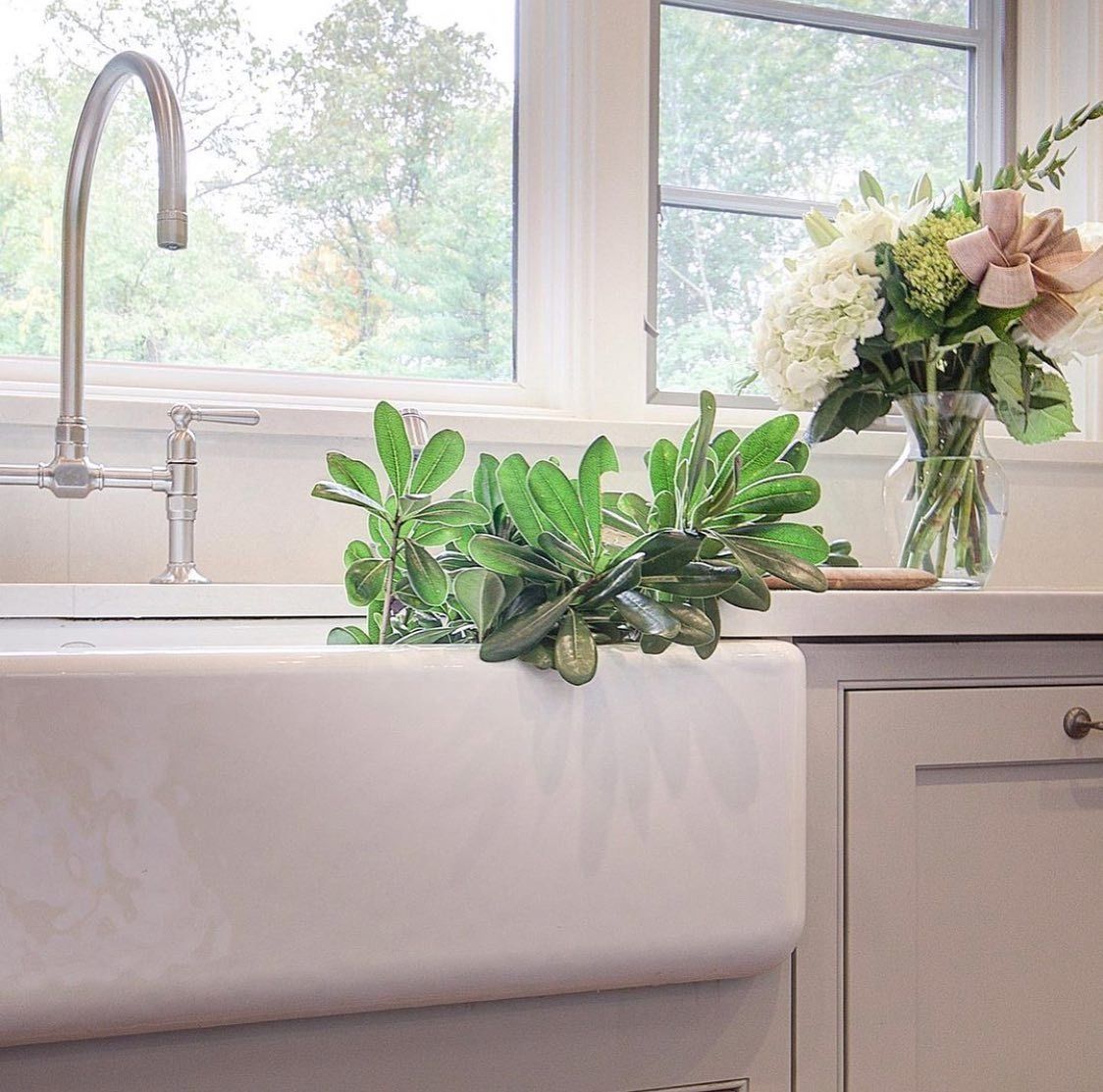 White farmhouse sink with faucet, greenery, and flowers in front of a window.