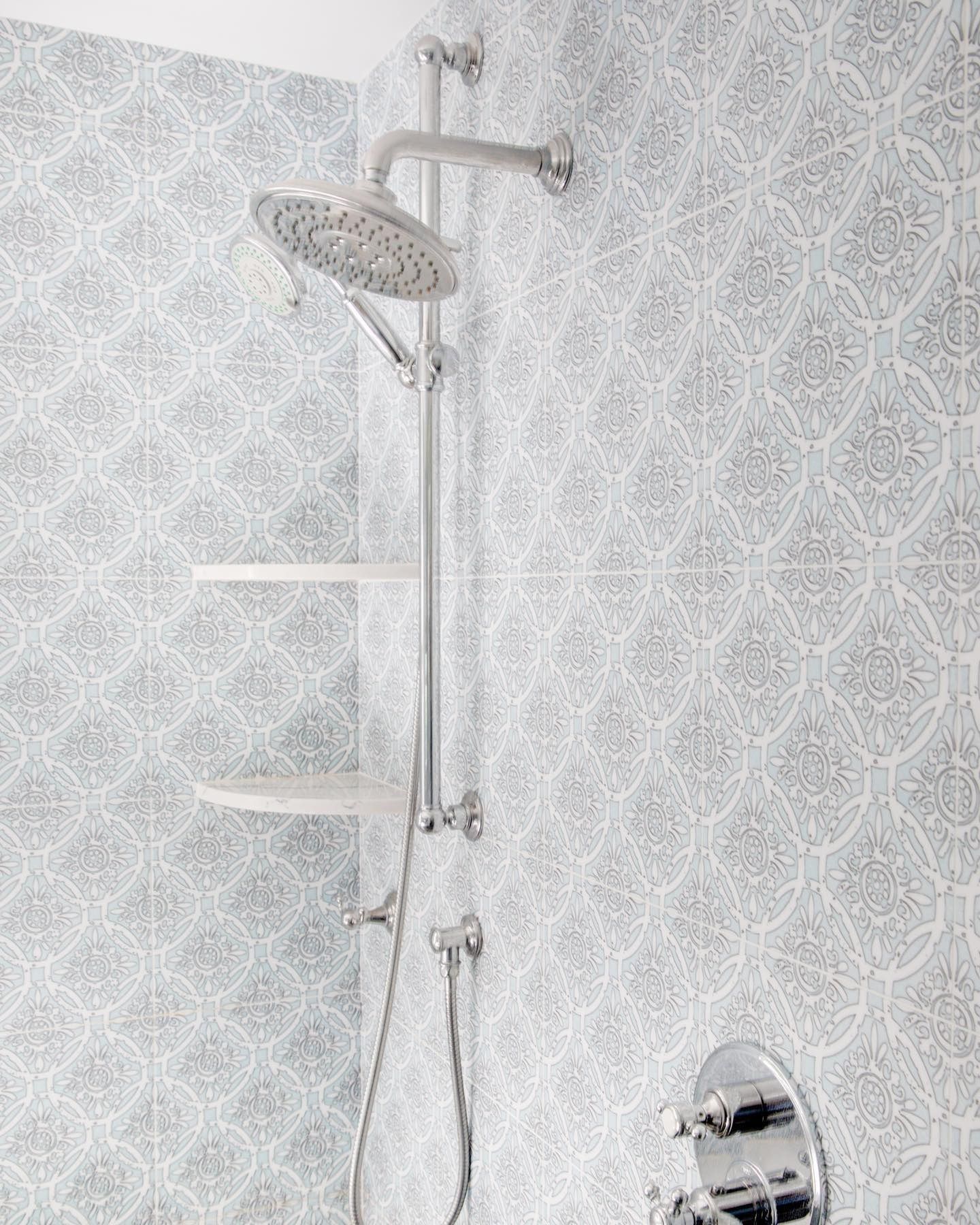 Shower fixture on patterned tile wall, with two shelves and a handheld showerhead.