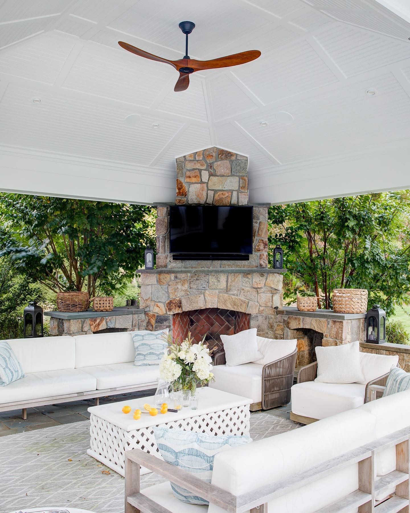 Outdoor living space with white sofas, stone fireplace, and ceiling fan.