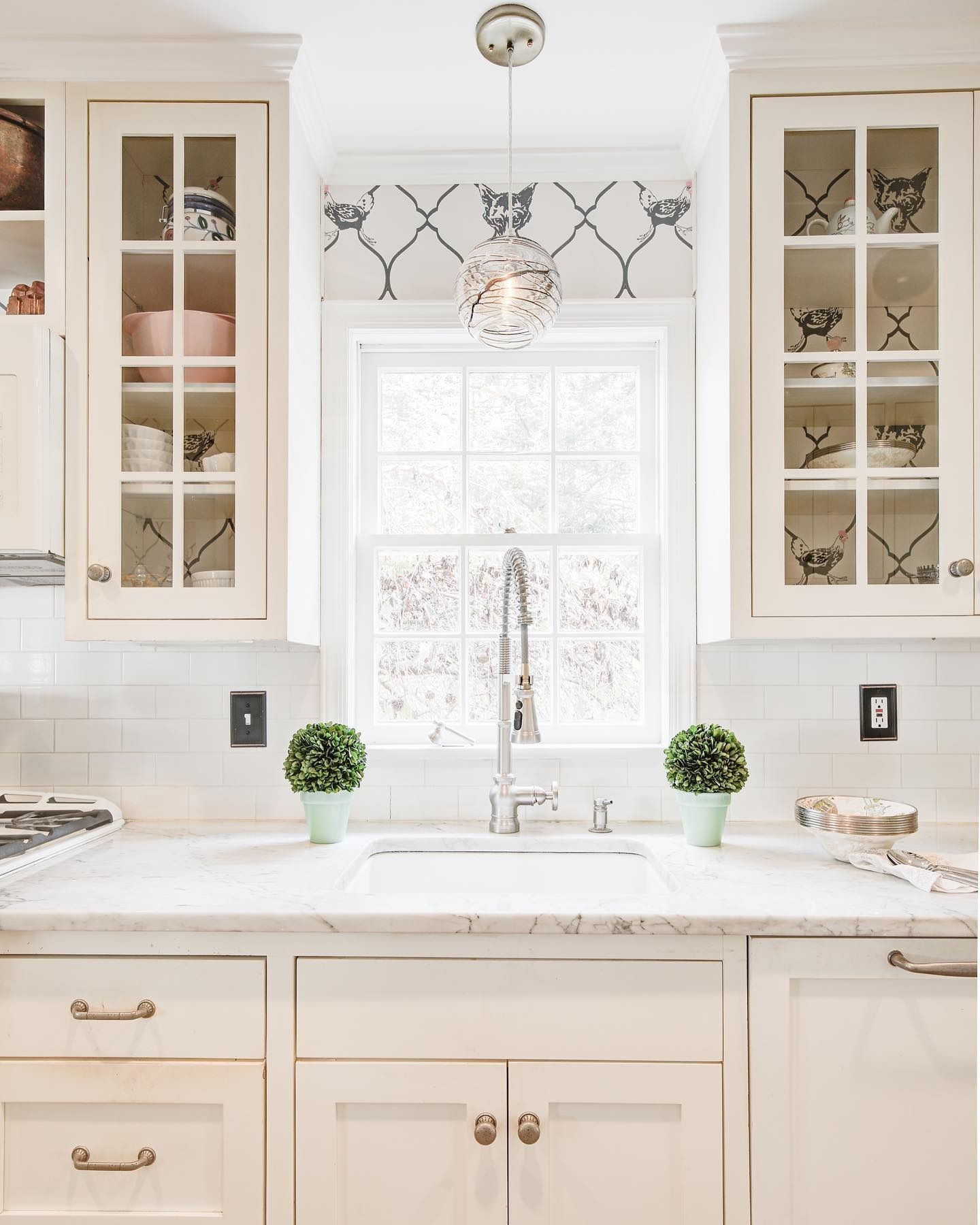 White kitchen with window over sink.  Cabinets and countertop. Two small plants.  Bird-patterned valance.