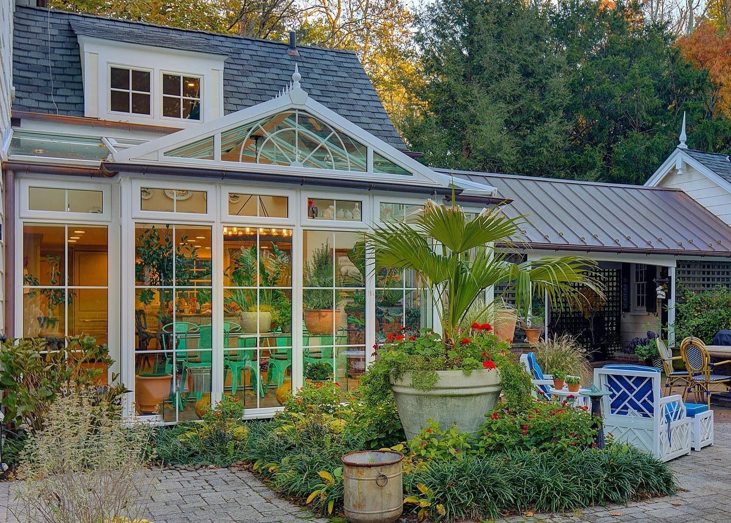 Greenhouse attached to a white building; plants and patio furniture are in the yard.