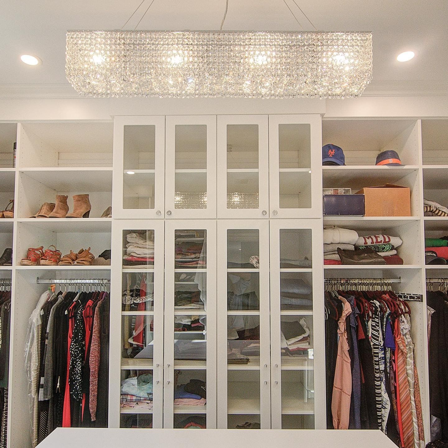 Walk-in closet with white shelves and cabinets, hanging clothes, shoes, and a crystal chandelier.