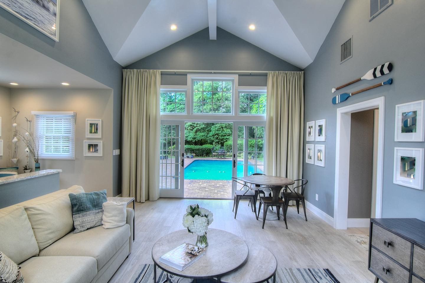 Living room with a pool view. Light blue walls, cream curtains, white sofa, round table, and framed pictures.