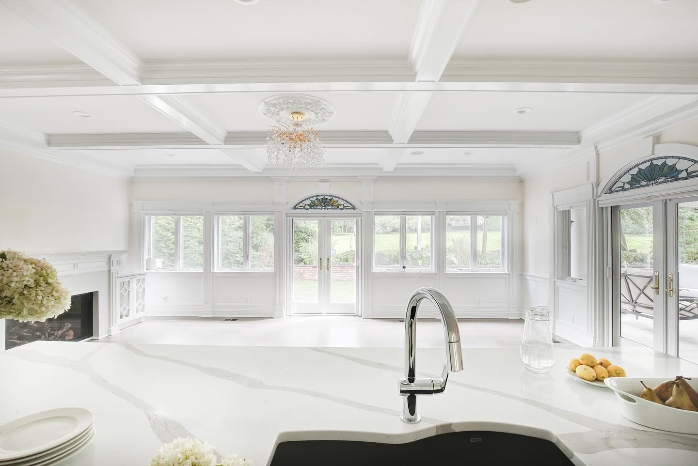 White kitchen with large windows, fireplace, and chandelier; bright, airy interior.