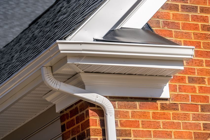 White gutter system on a red brick building with dark roof shingles. A downspout extends downward from the gutter.