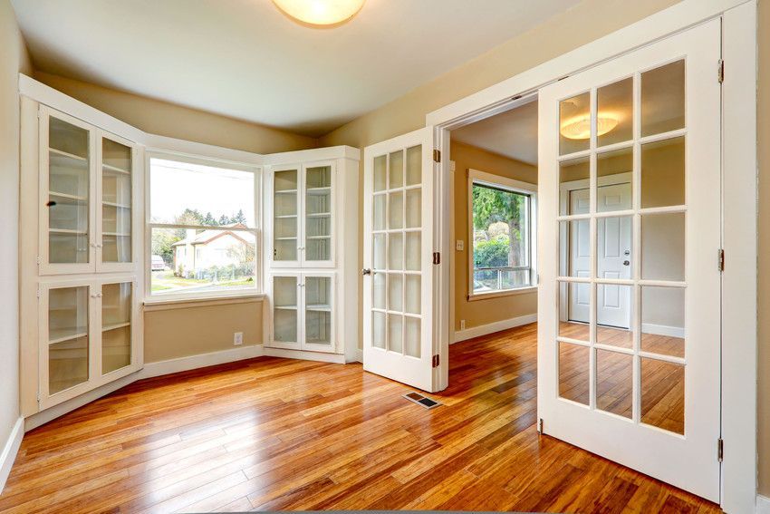 A light-filled room with hardwood floors, built-in white cabinets, a bay window, and French doors leading to another room.
