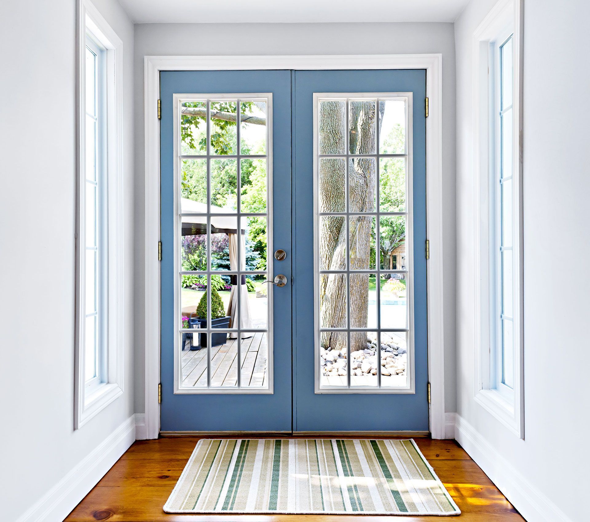 Blue double doors with glass panels, flanked by white-framed windows. A striped rug rests on a wooden floor in the entryway.