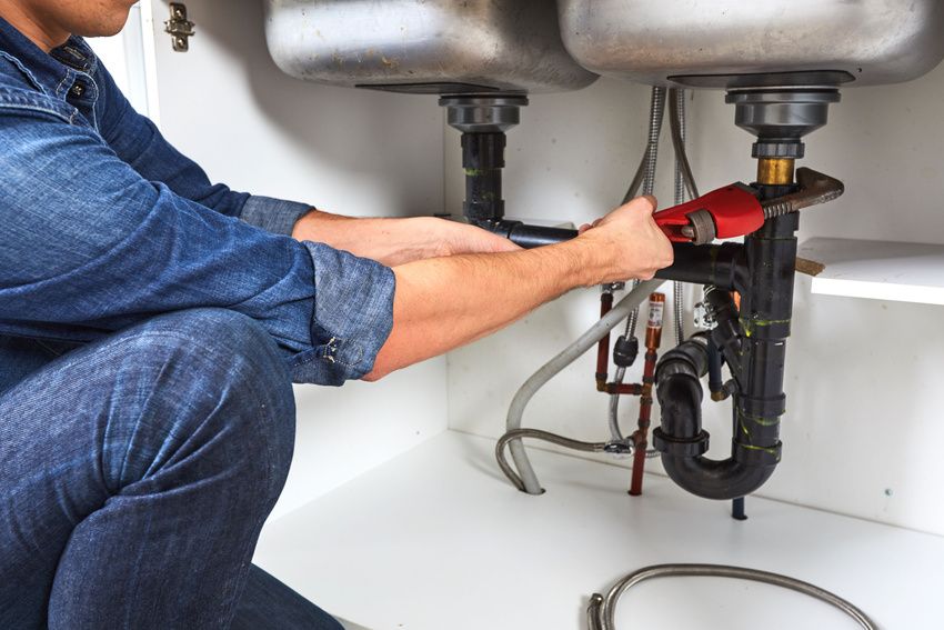A person in a denim shirt uses a wrench to repair plumbing under a stainless steel sink.