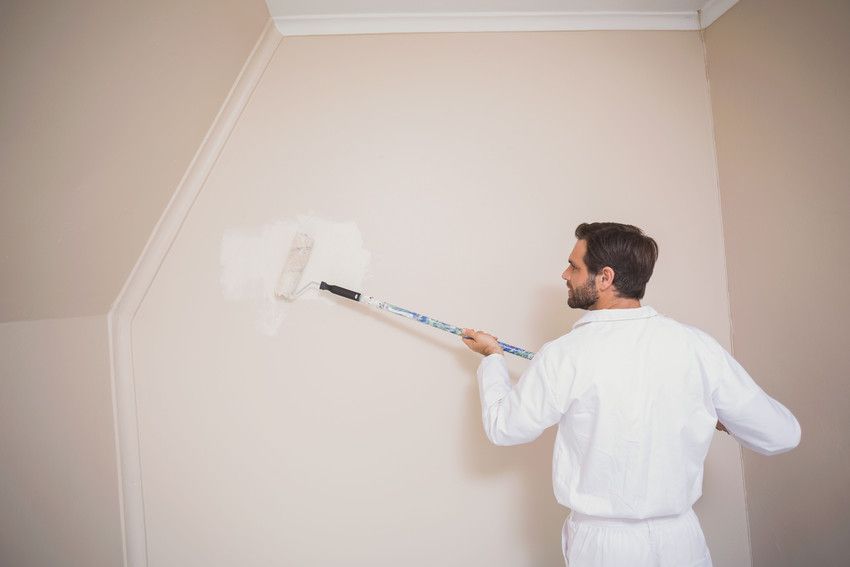 Man in white coveralls painting a wall with a roller; interior setting.