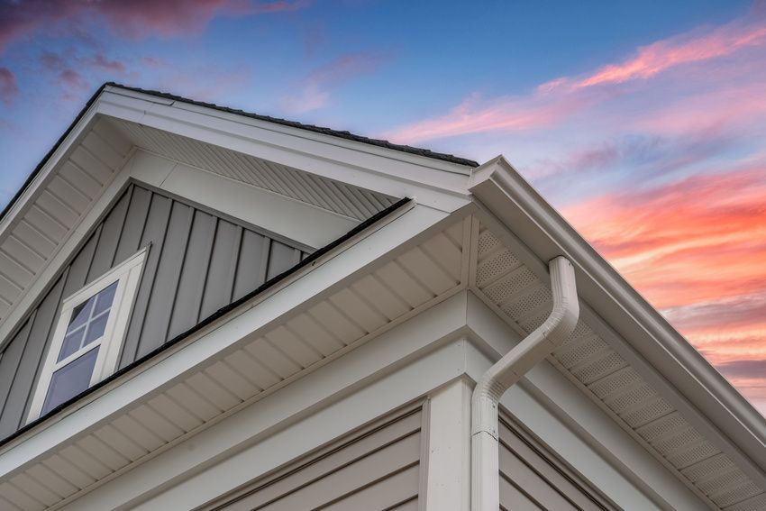 Close-up of a house exterior with a white trim, gray siding, and a gutter against a sunset sky.
