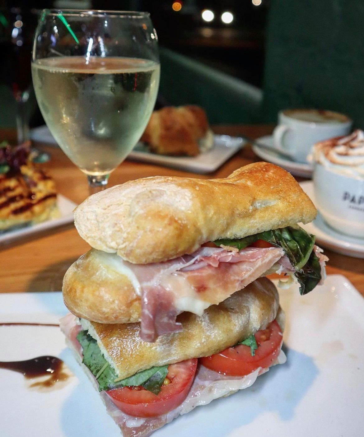 Sandwich with prosciutto and tomato, white wine, and other food items on a table.