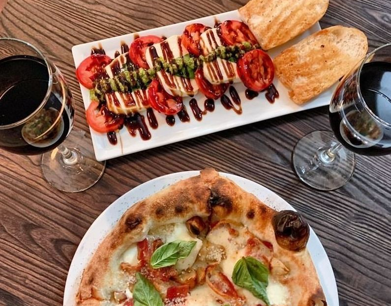 Italian meal: pizza, Caprese salad, toasted bread, red wine glasses on table.