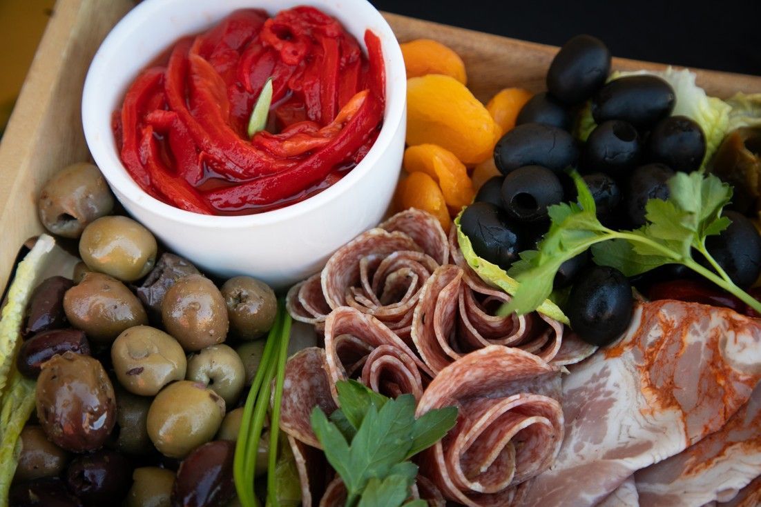 A wooden tray filled with meats , olives , peppers and a bowl of sauce.
