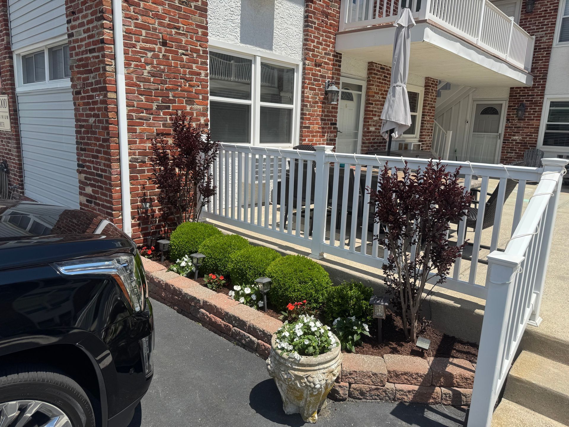 Brick townhouse entry with white ramp, railings, potted plant, and flower bed beside a parked car