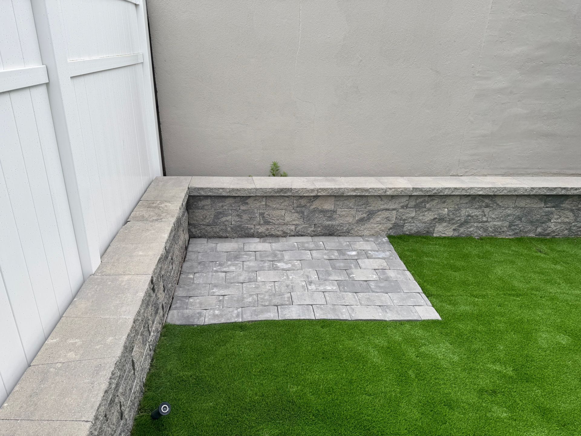 Small backyard with gray stone patio, raised planter bed, white fence, and green artificial grass.