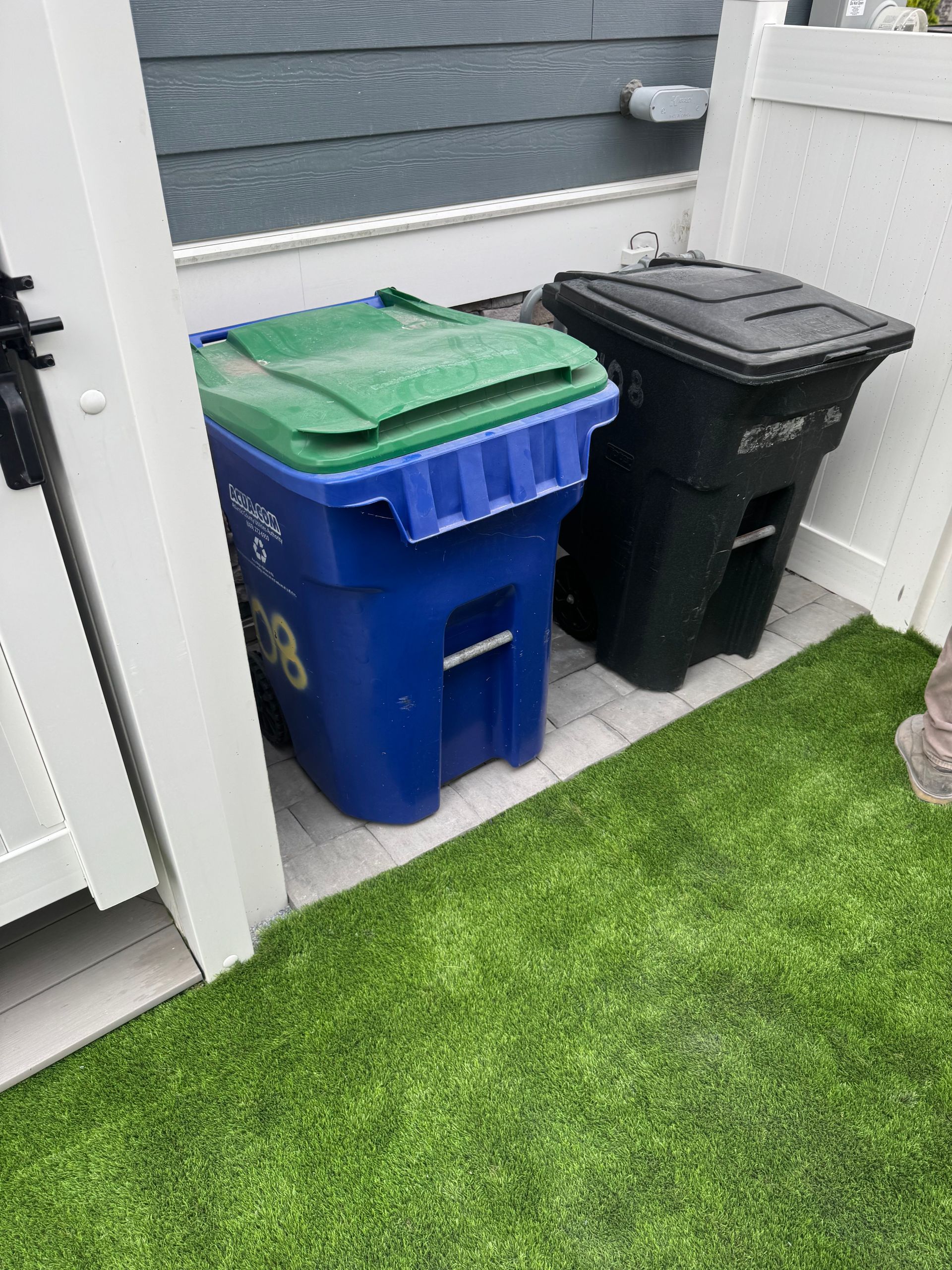 Blue and black wheeled trash bins beside a white wall on a narrow patio next to green grass.