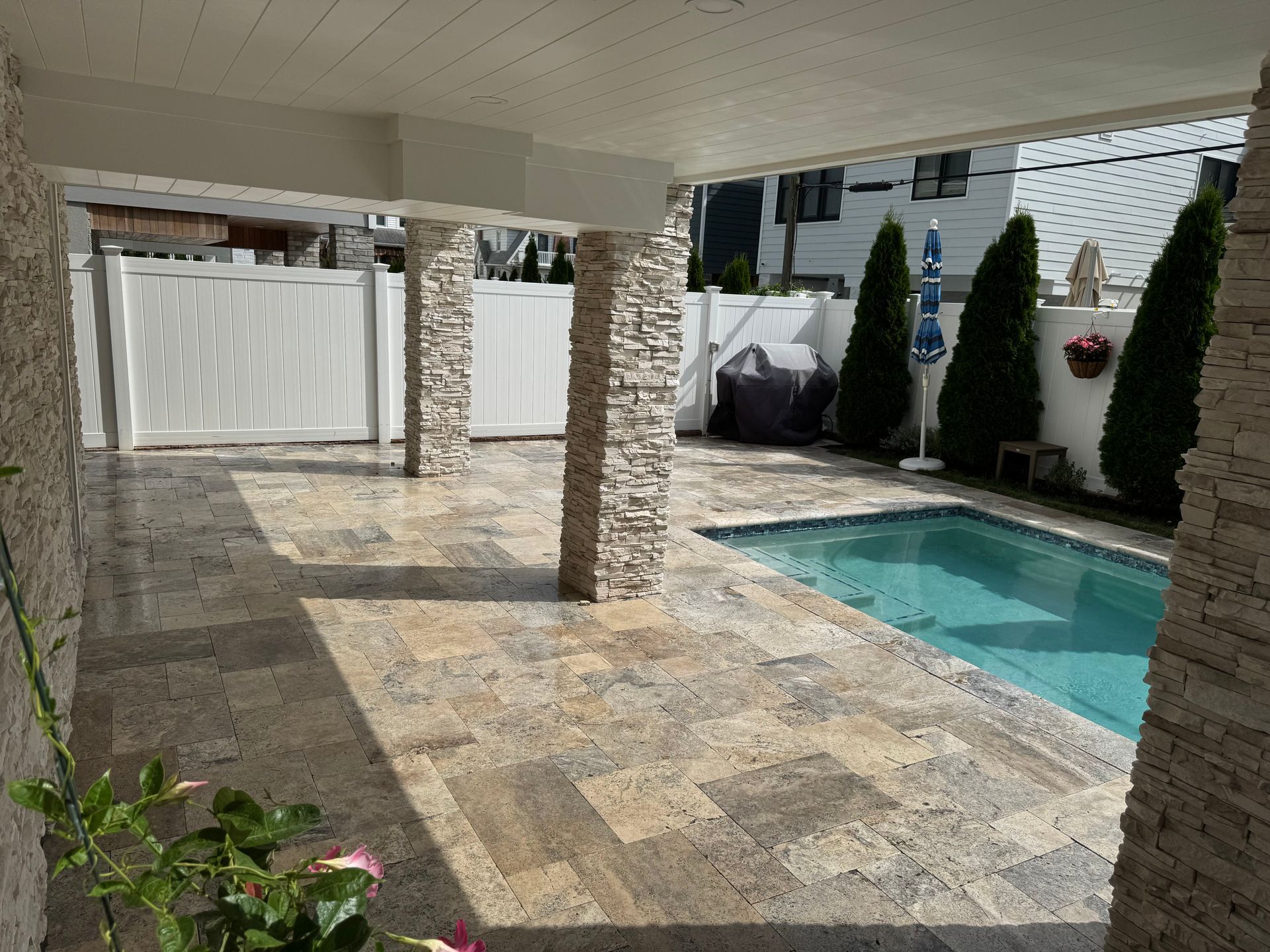 Covered patio beside a blue swimming pool with stone columns and white fencing