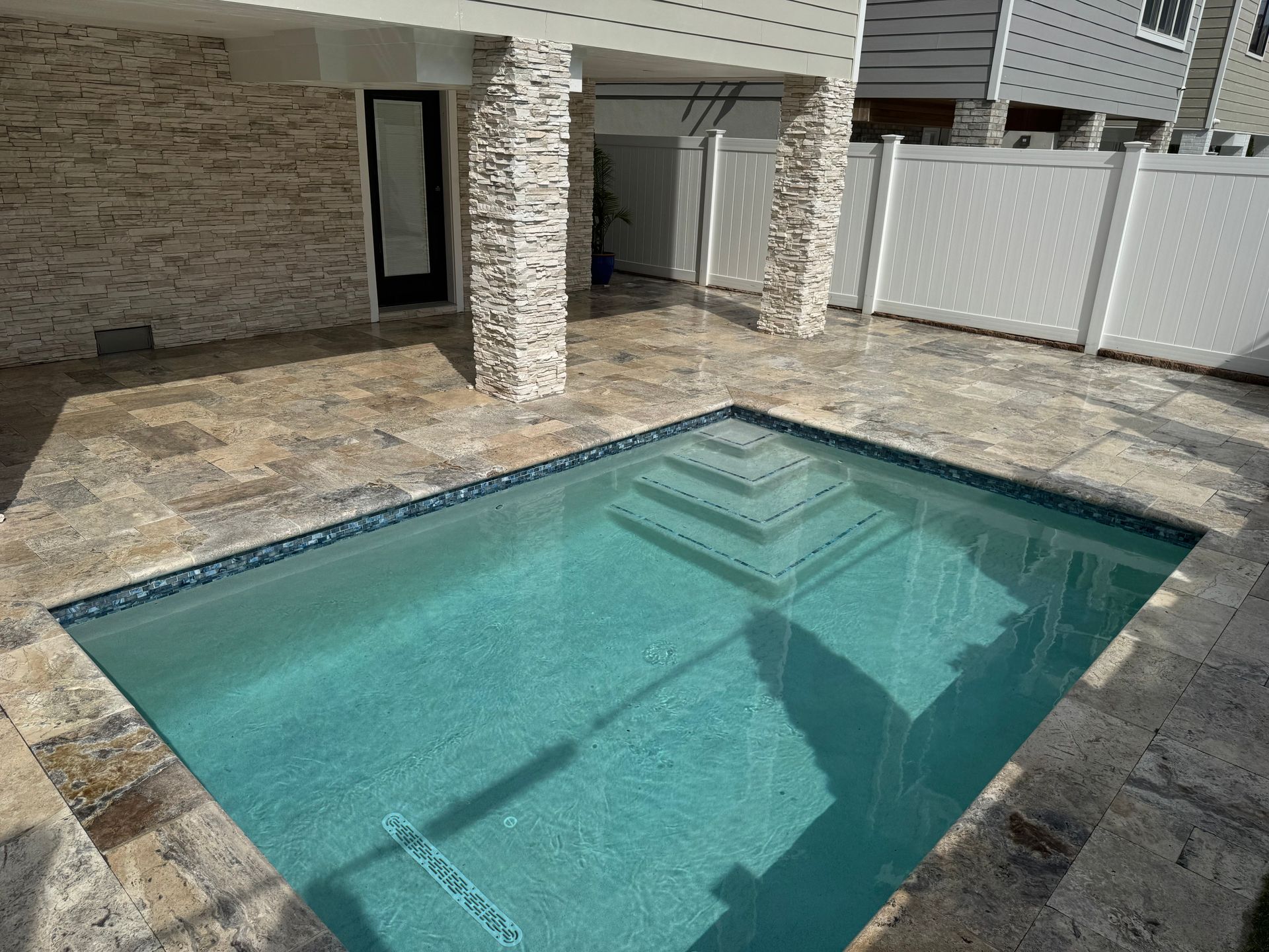 Backyard patio with a small rectangular swimming pool and stone paving beside a house and white fence