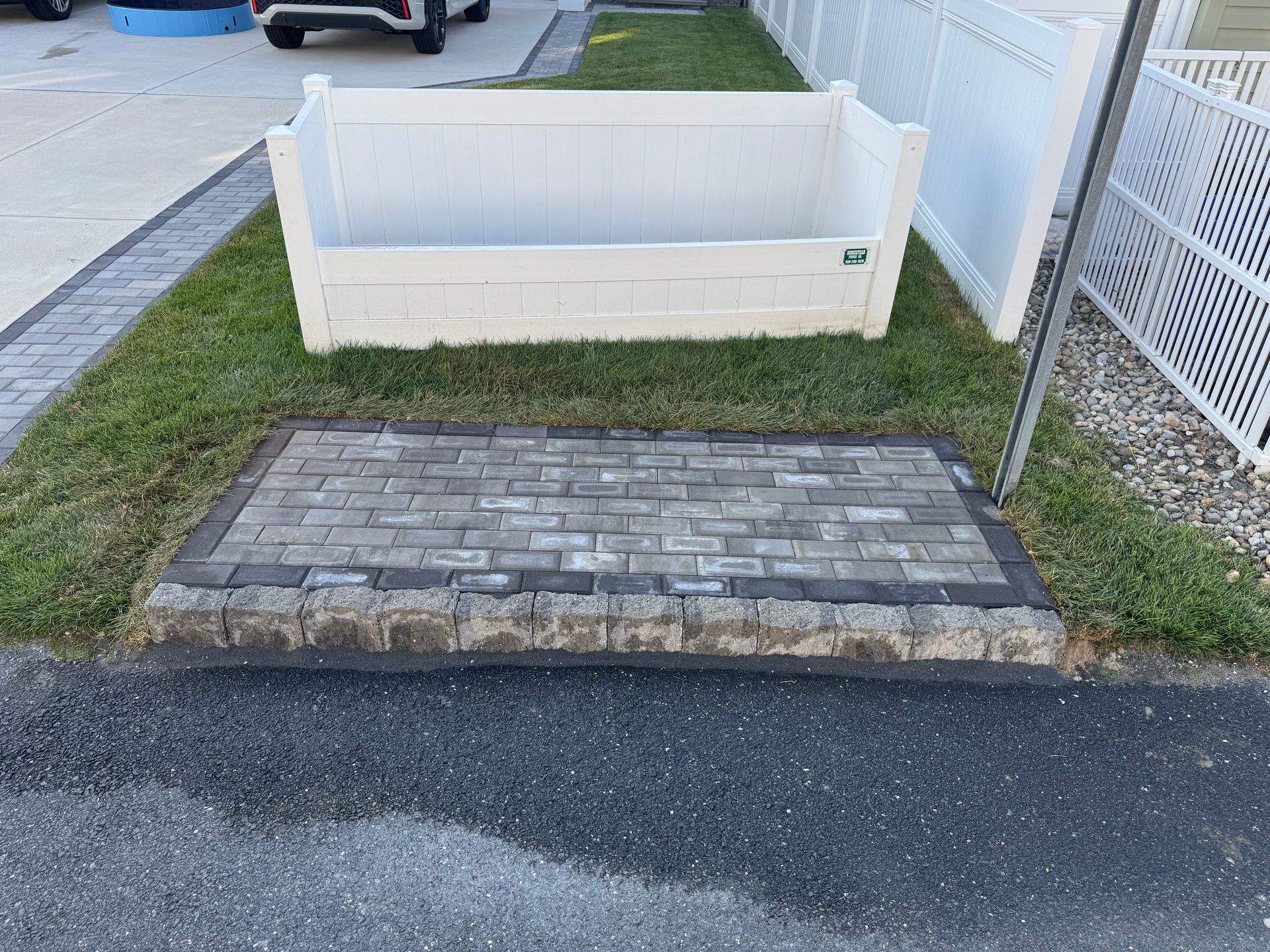 Brick-paved curb ramp beside a grassy strip and white planter along a sidewalk near a driveway