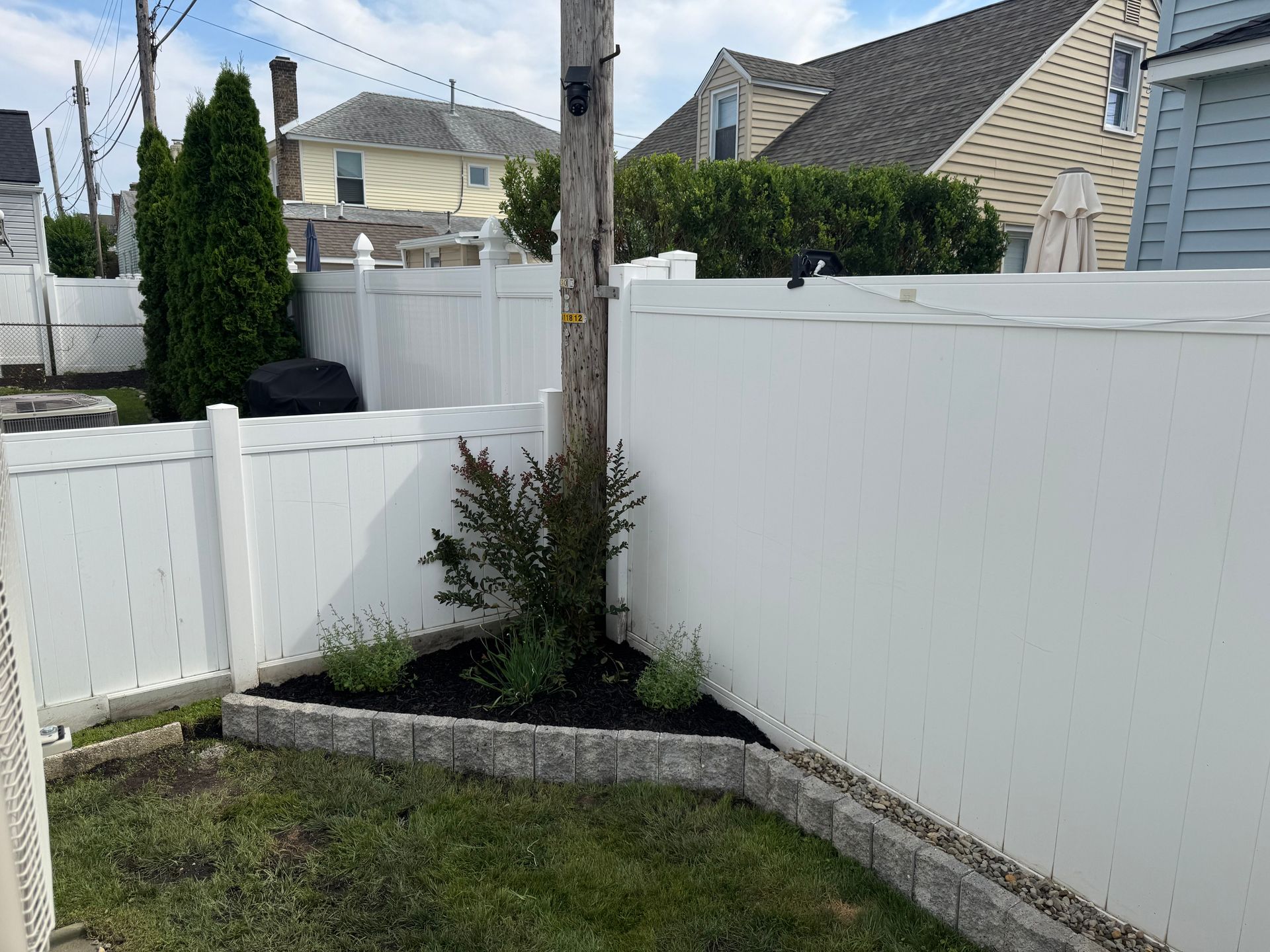 Small backyard corner with white fence, raised stone bed, and a young tree.