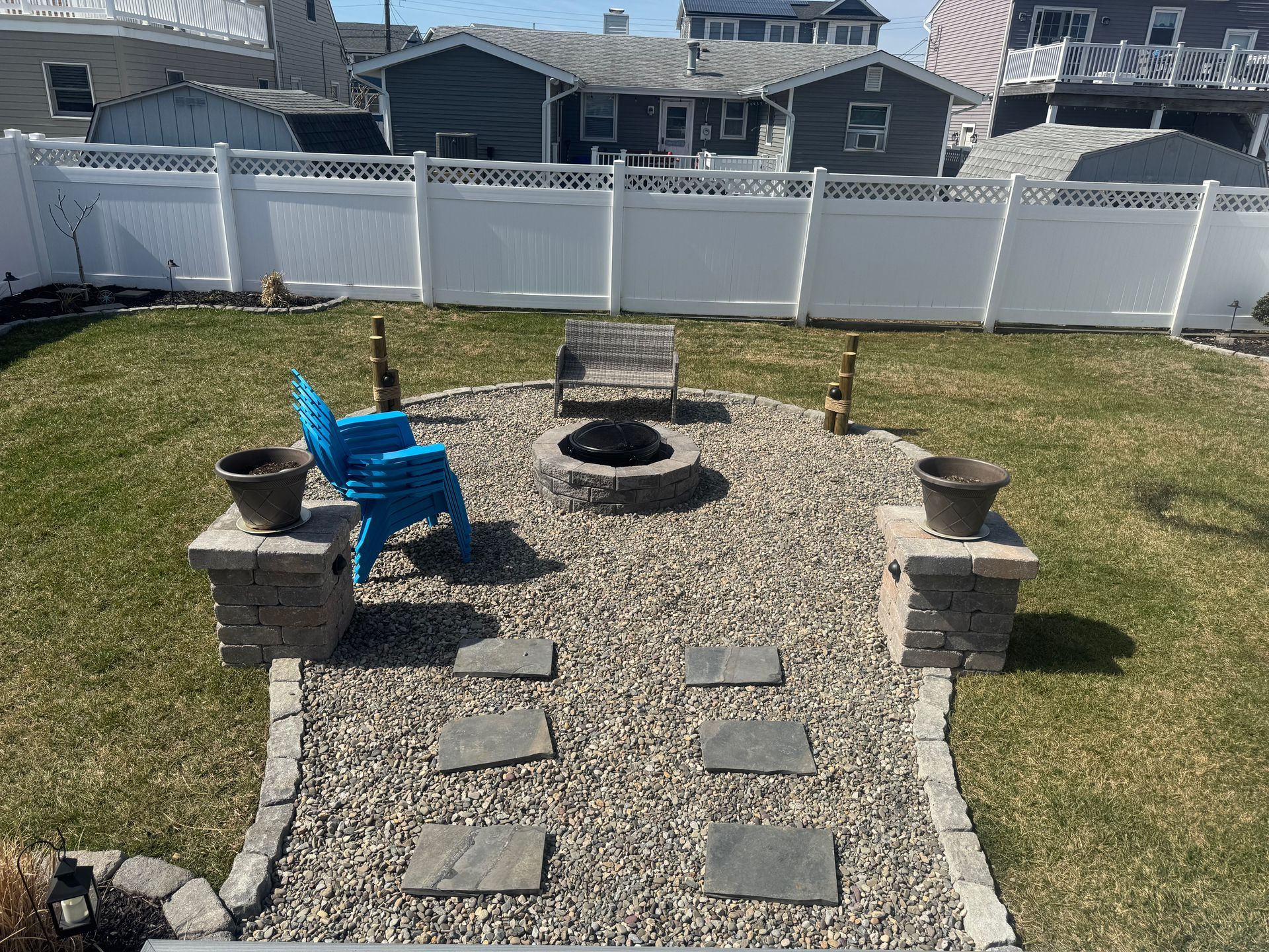 Backyard fire pit area with stone pathway, blue chair, and white fence.