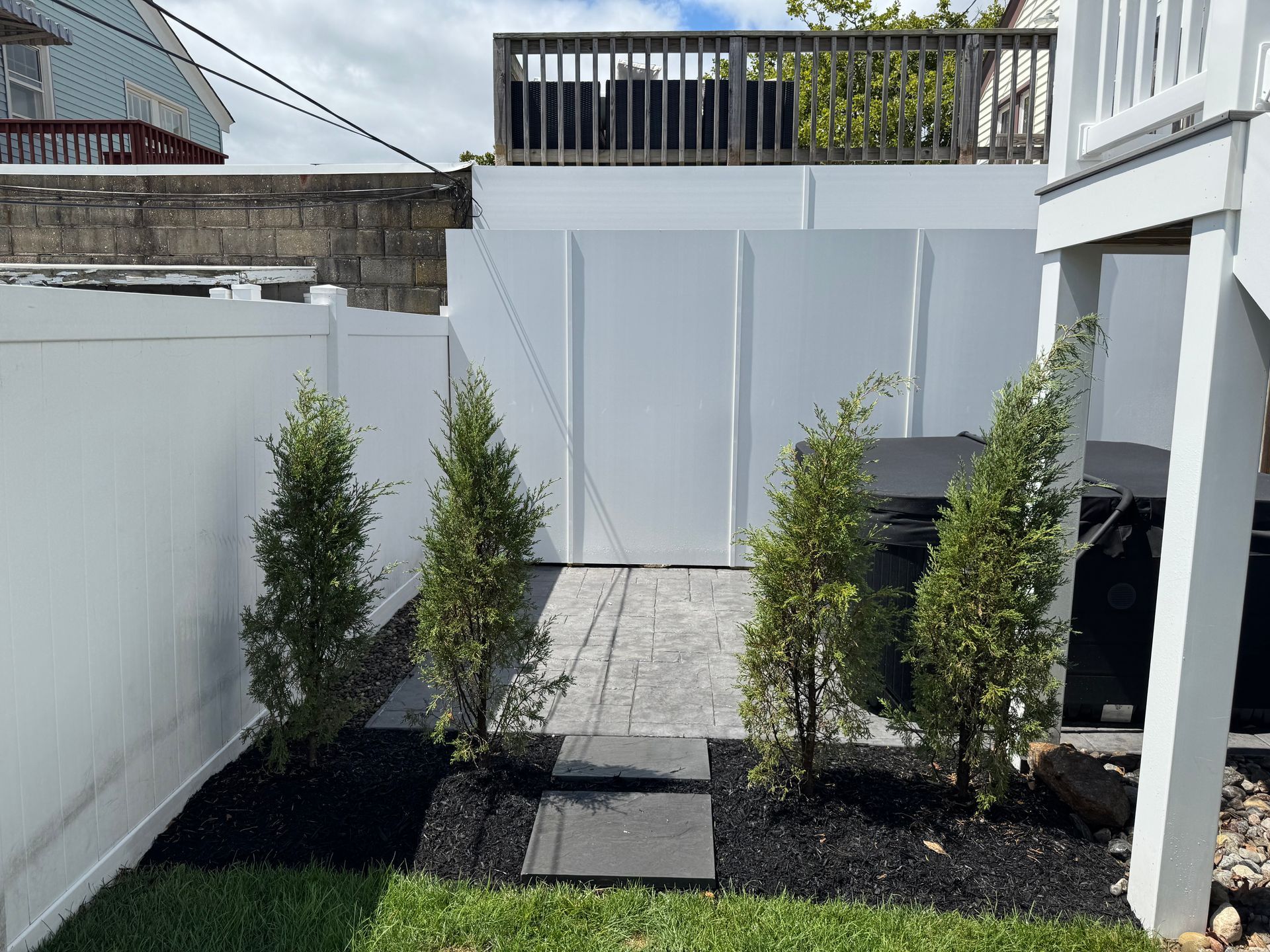 White fenced-in backyard area with four small trees and dark mulch. A deck and stone path are also visible.