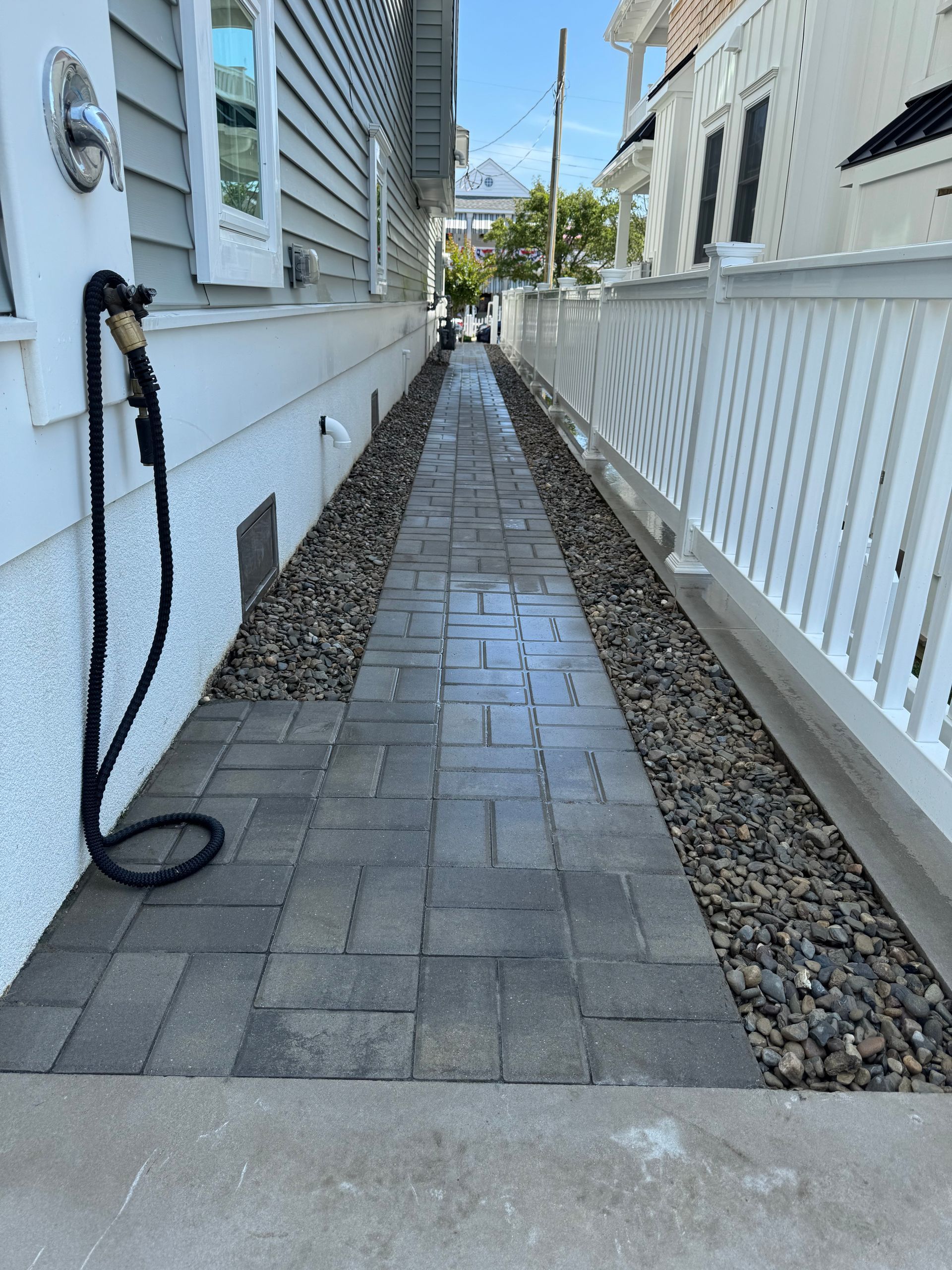 Brick pathway between two buildings, bordered by pebbles. A water hose hangs on the left.