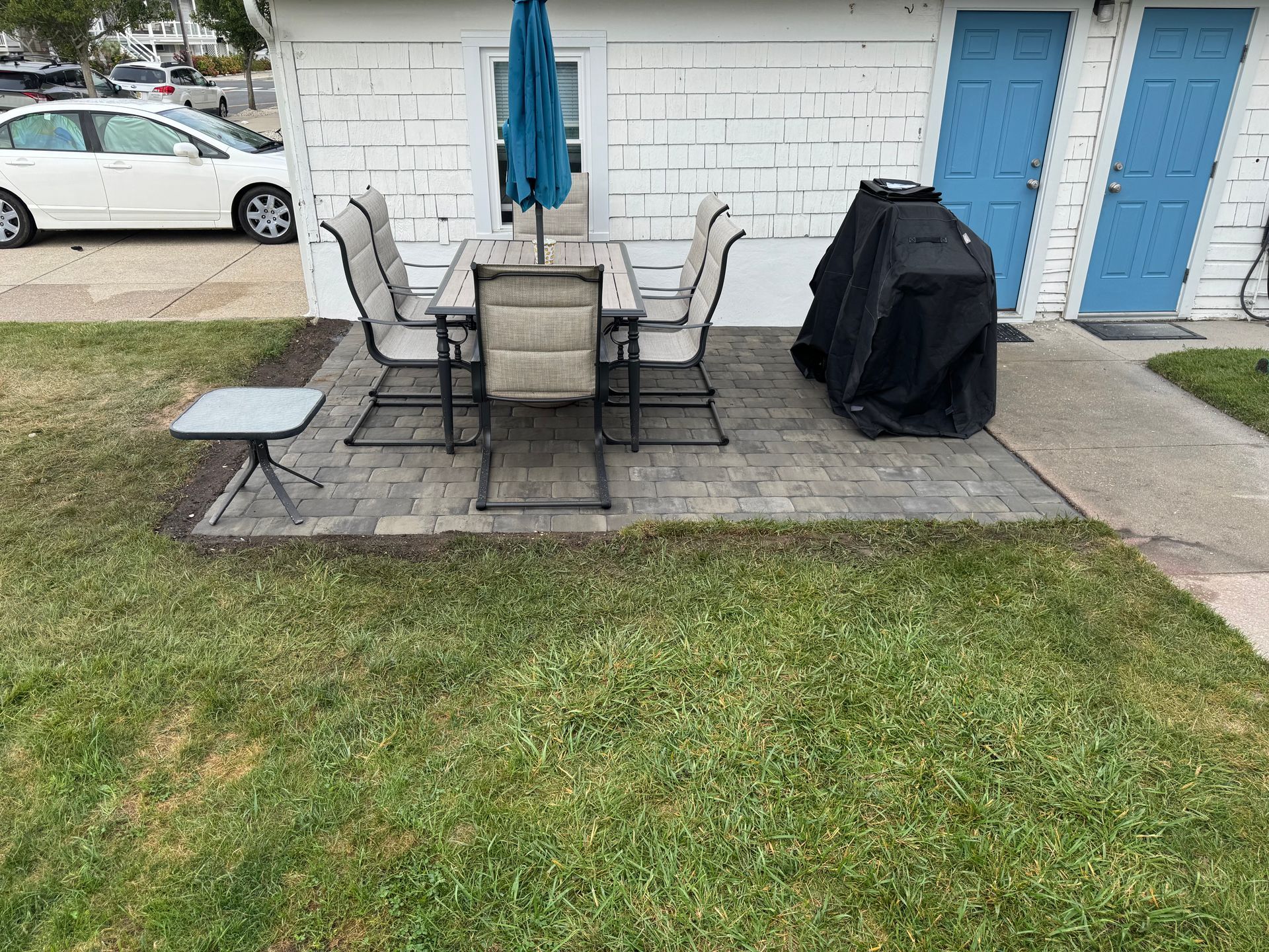 Patio with table, chairs, umbrella, grill, and small side table on a brick surface; lawn in front.
