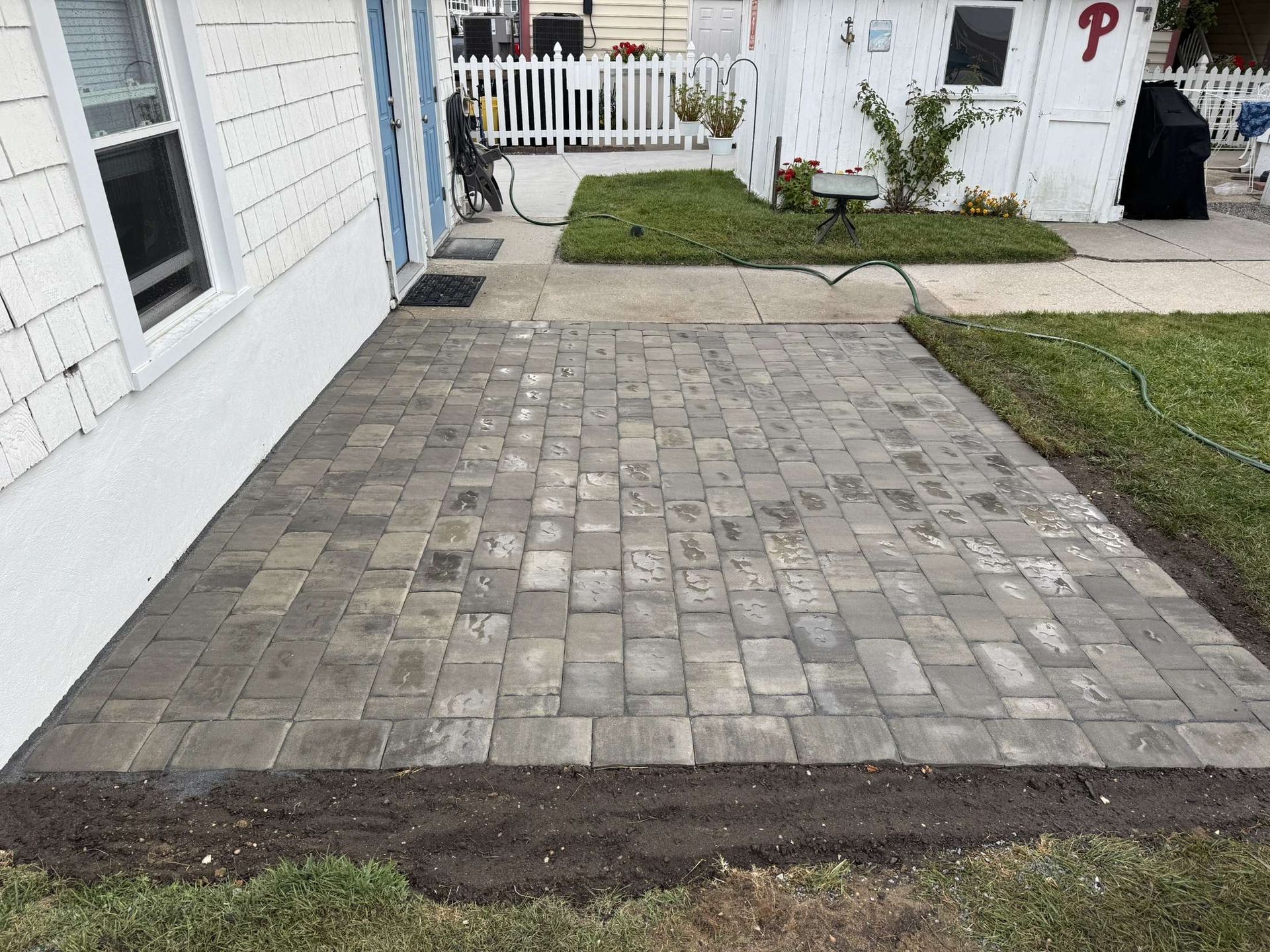 Paved patio next to a house with a white shed in the background. Grass and flower beds are adjacent.